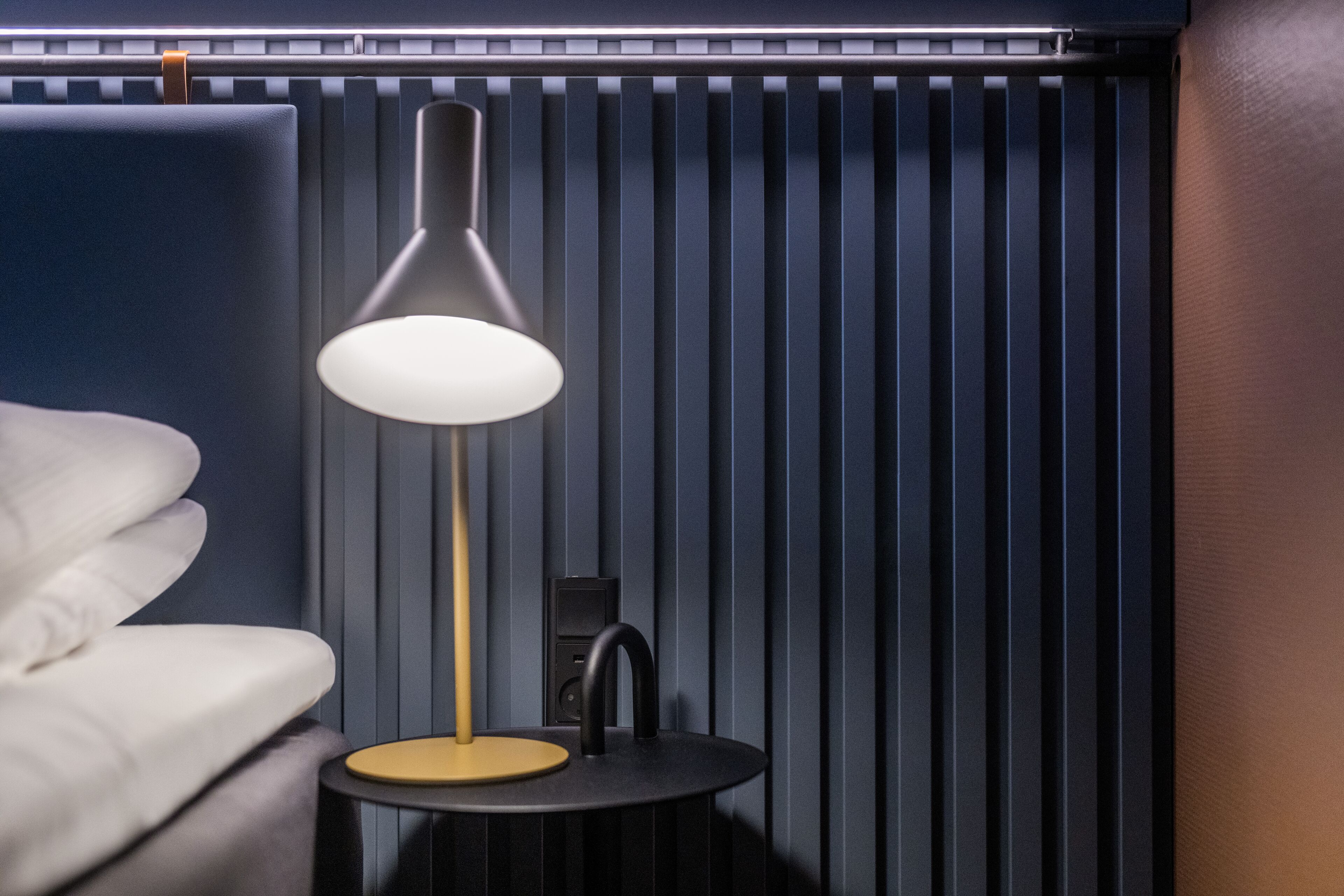 En moderne lampe på et lille bord ved siden af en hvid stol, med lodrette skodder i baggrunden.