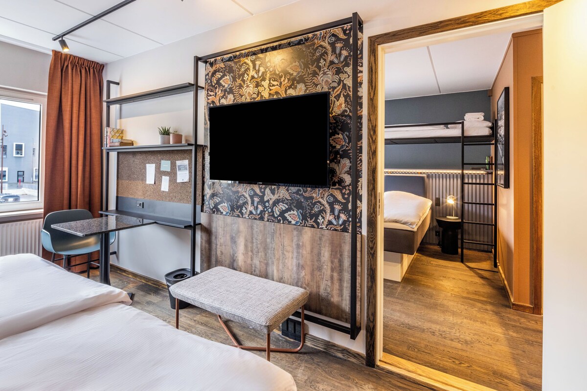 A hotel room with a bed, a desk, a chair, a TV, and a bunk bed.