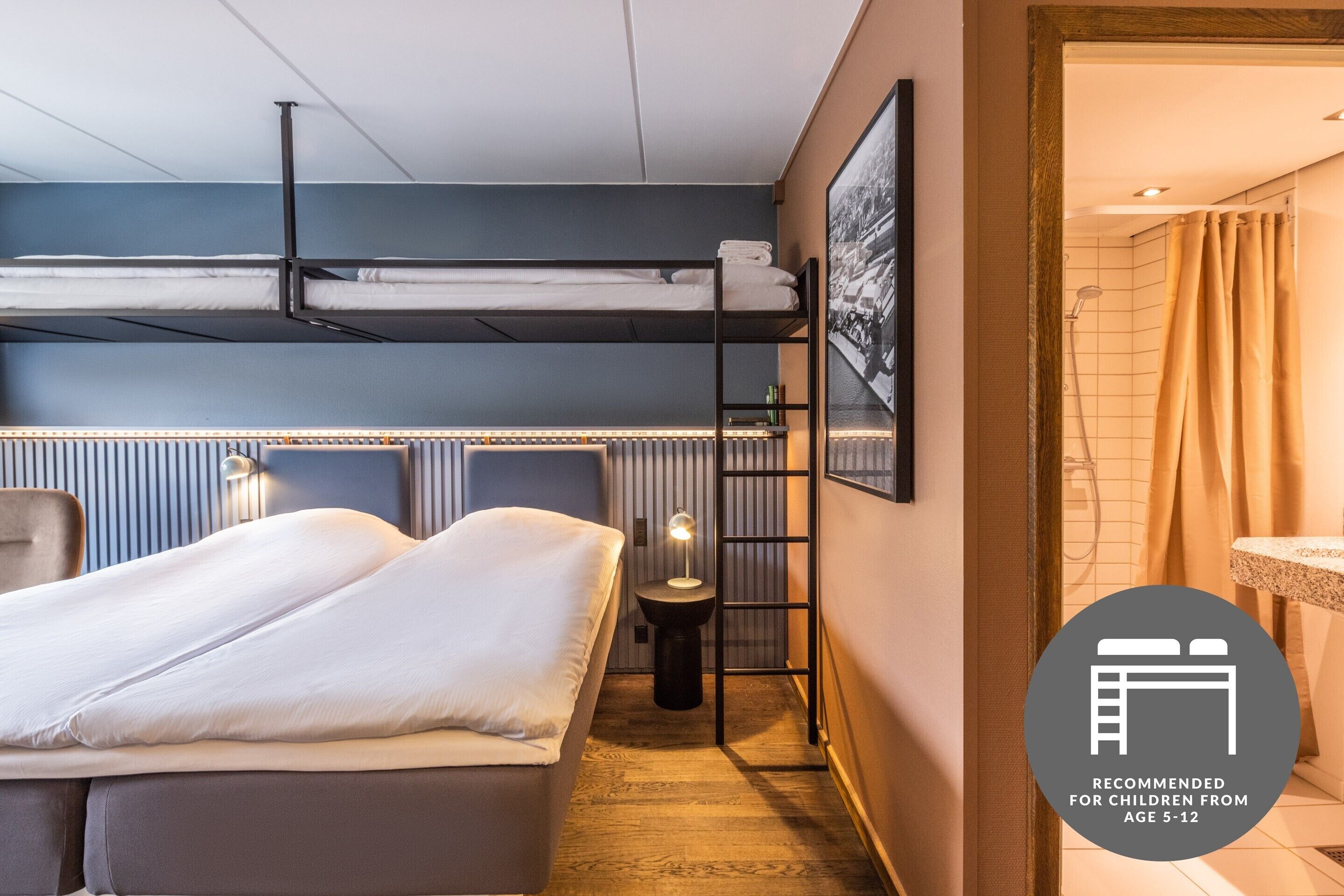 Habitación de hotel con una cama nido, una mesita de noche con lámpara, un taburete pequeño y un rincón de ducha.