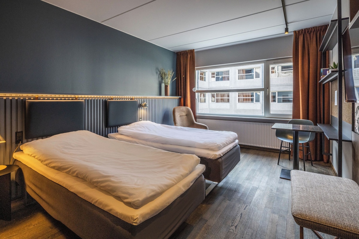 A hotel room with two beds, a chair, a table, and a view of buildings through the window.