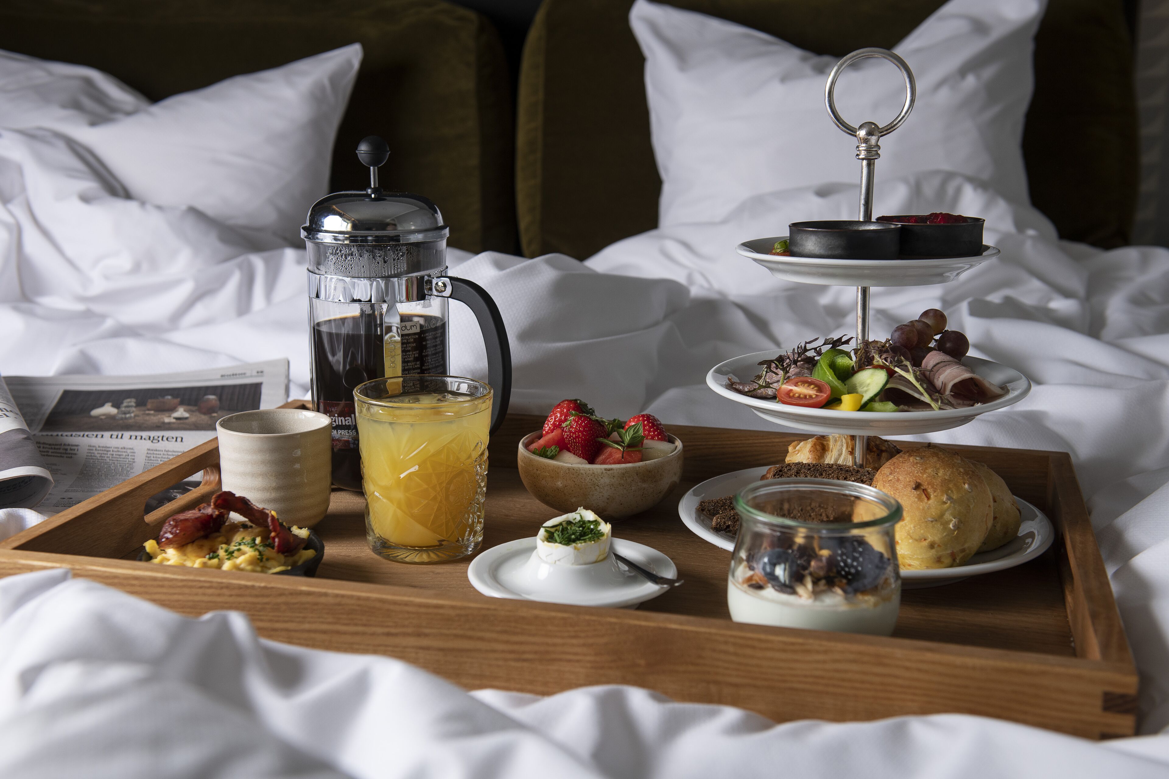 A breakfast tray on a bed with a coffee maker, a glass of orange juice, a bowl of strawberries, a plate of pastries, and a small jar of yogurt.