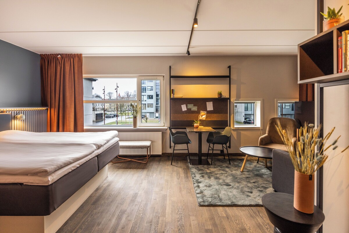 A modern hotel room with a bed, a desk, a chair, a small table, a radiator, and a view of the outside.