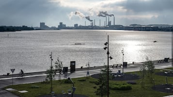 A waterfront view with a road, greenery, and industrial buildings in the background.