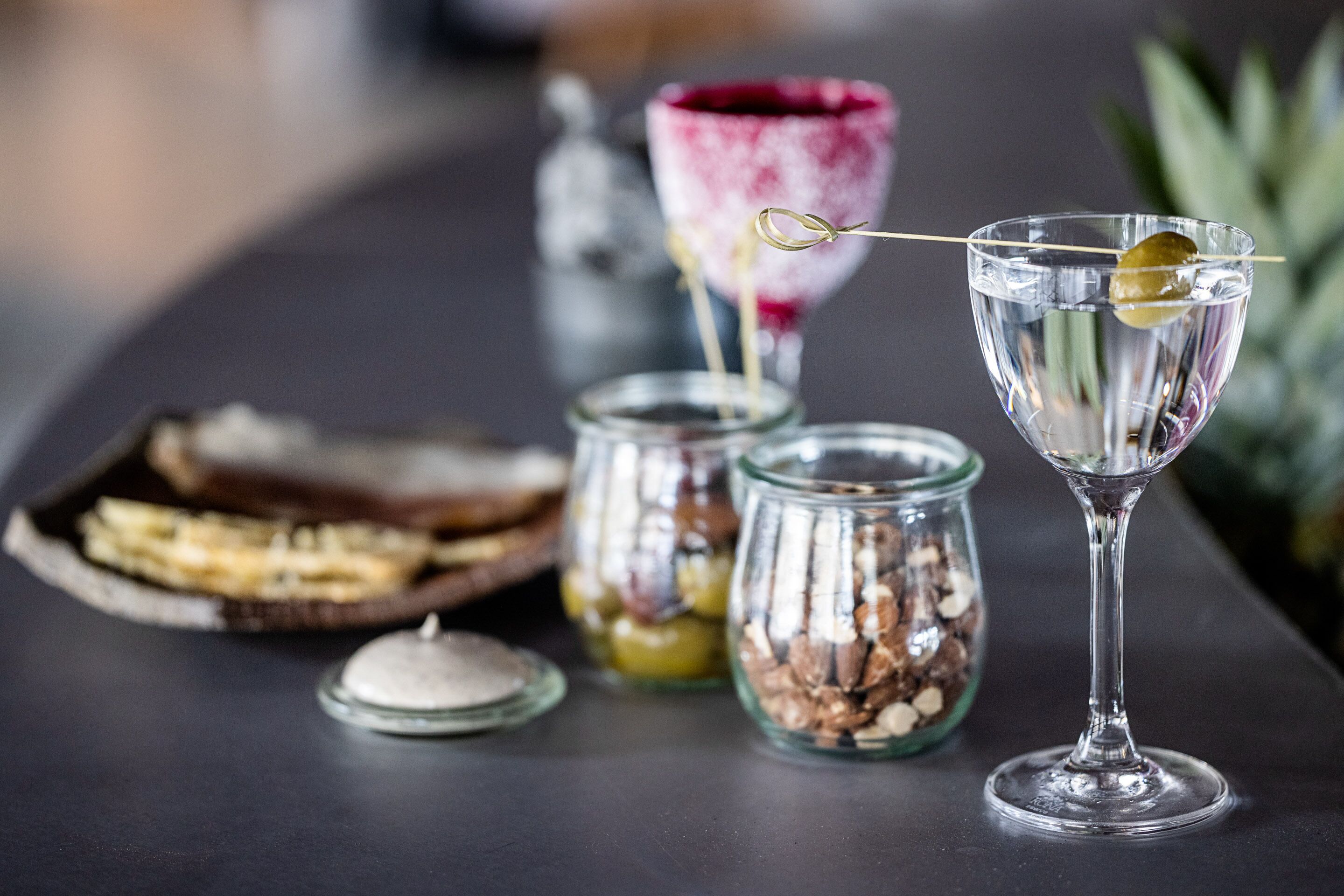 A bar setting with a glass of water, a martini glass with an olive, and two jars of nuts and dried fruits.