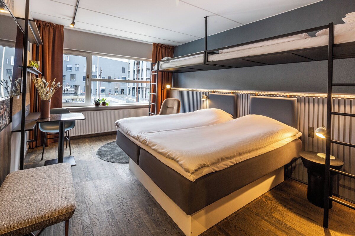 A hotel room with a bunk bed, a desk, a chair, a small table, and a view of the city through the window.