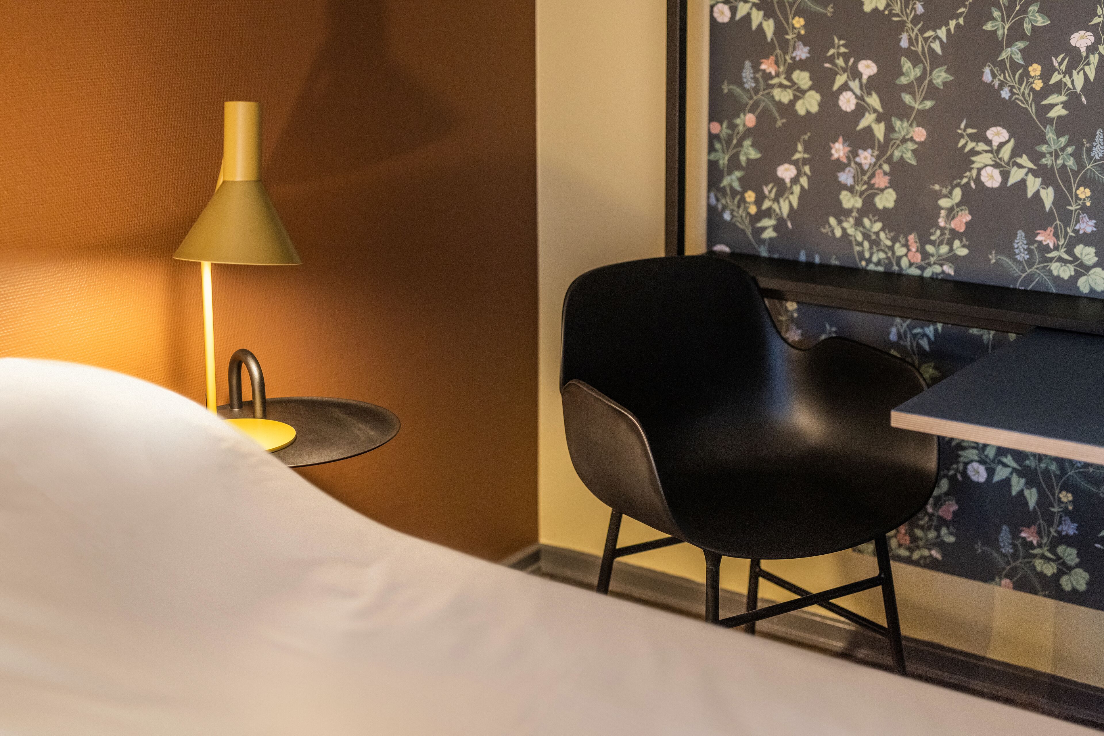 A hotel room with a bed, a desk, a chair, a lamp, and a floral wallpaper.
