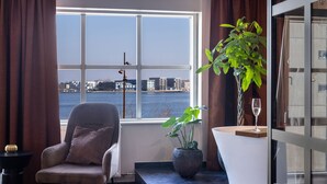 A modern living room with a view of a waterfront, a comfortable armchair, a potted plant, and a glass of wine on a white table.
