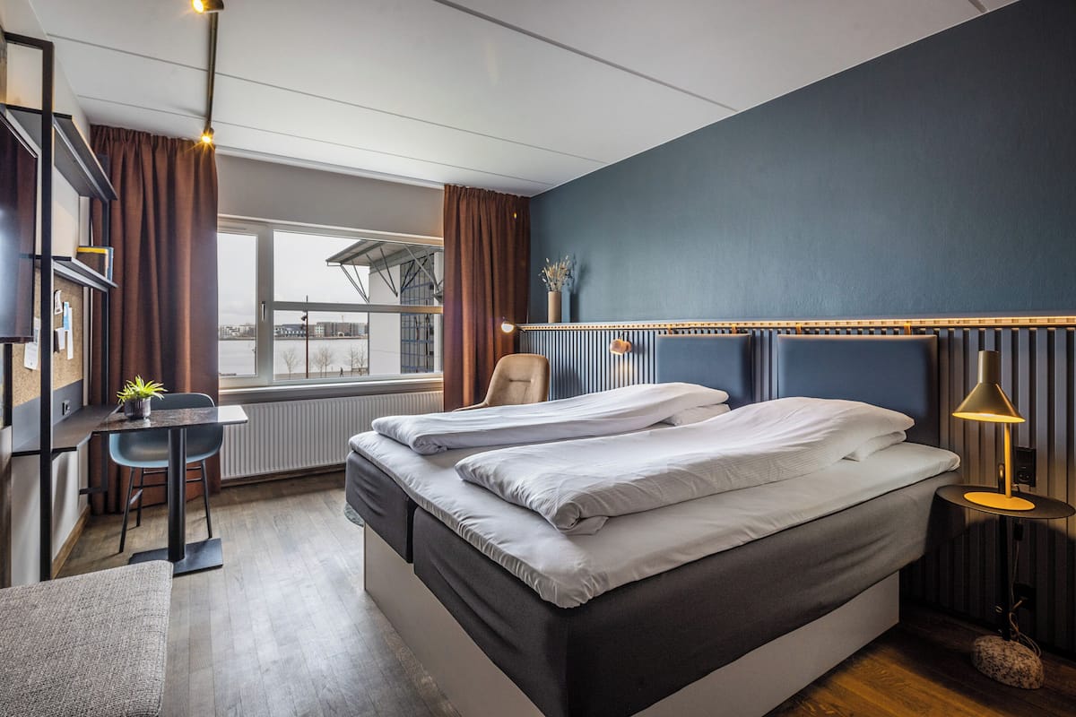 A double bed with a headboard, a small table with a chair, a desk with a chair, and a window with curtains.