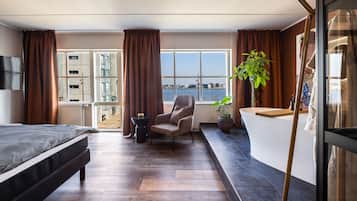 Una habitación de hotel moderna con cama, zona de estar, ventana grande con vista al agua y mostrador de recepción.