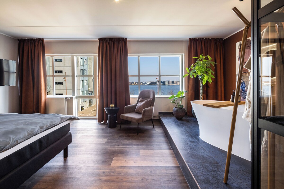 A modern hotel room with a bed, a seating area, a large window with a view of the water, and a reception desk.