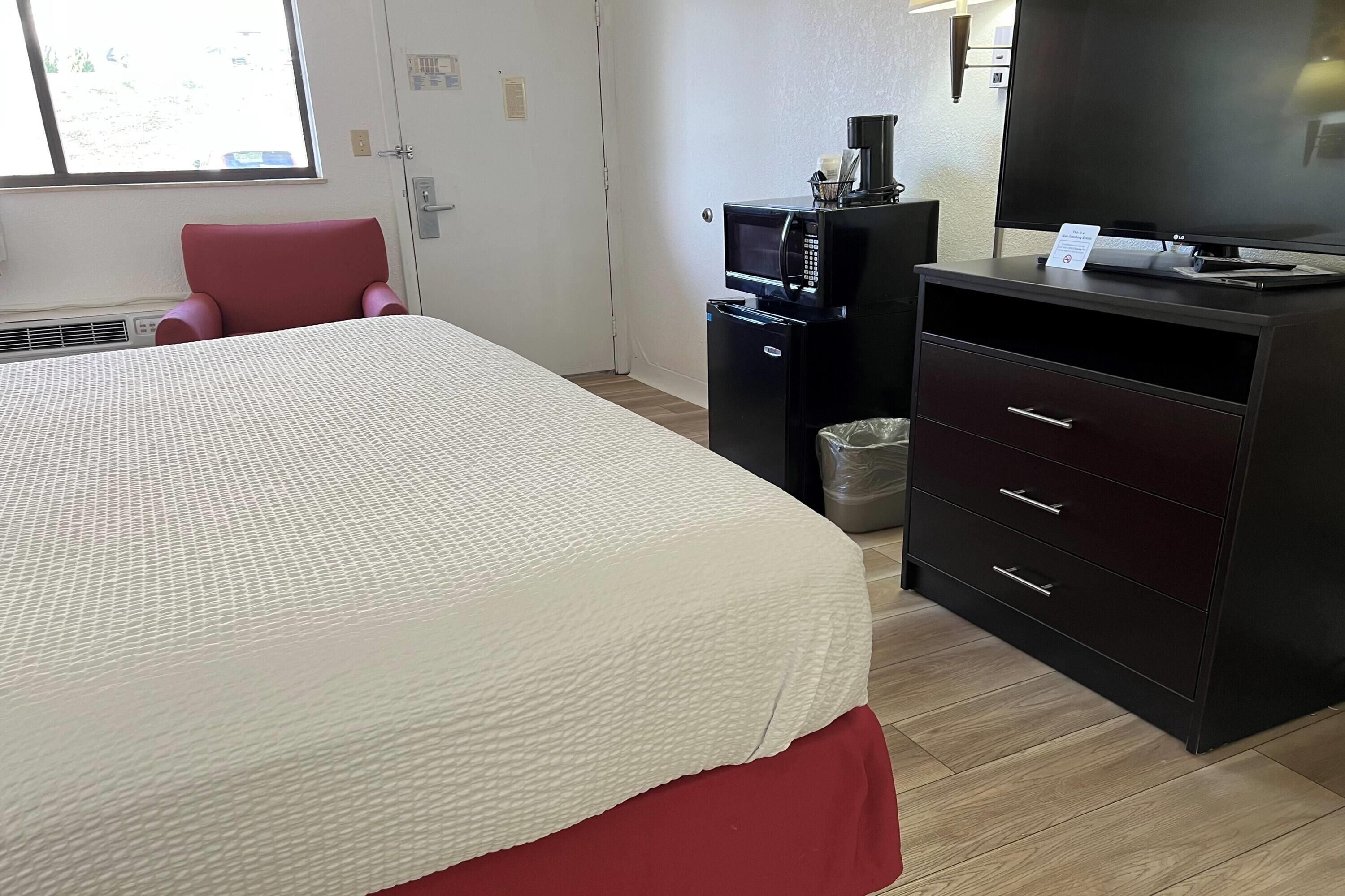 A hotel room with a large bed, a red chair, a desk with a computer and coffee maker, and a TV.