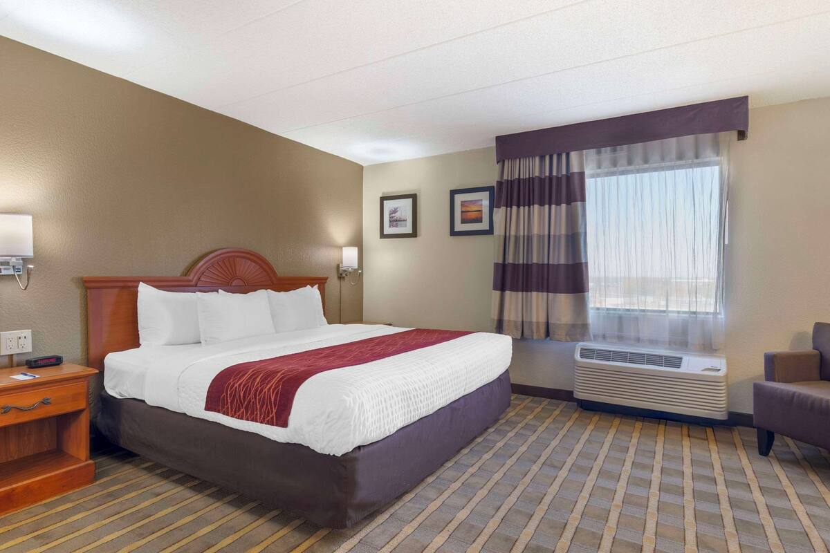 A hotel room with a bed, a nightstand, a chair, a window with curtains, and an air conditioning unit.