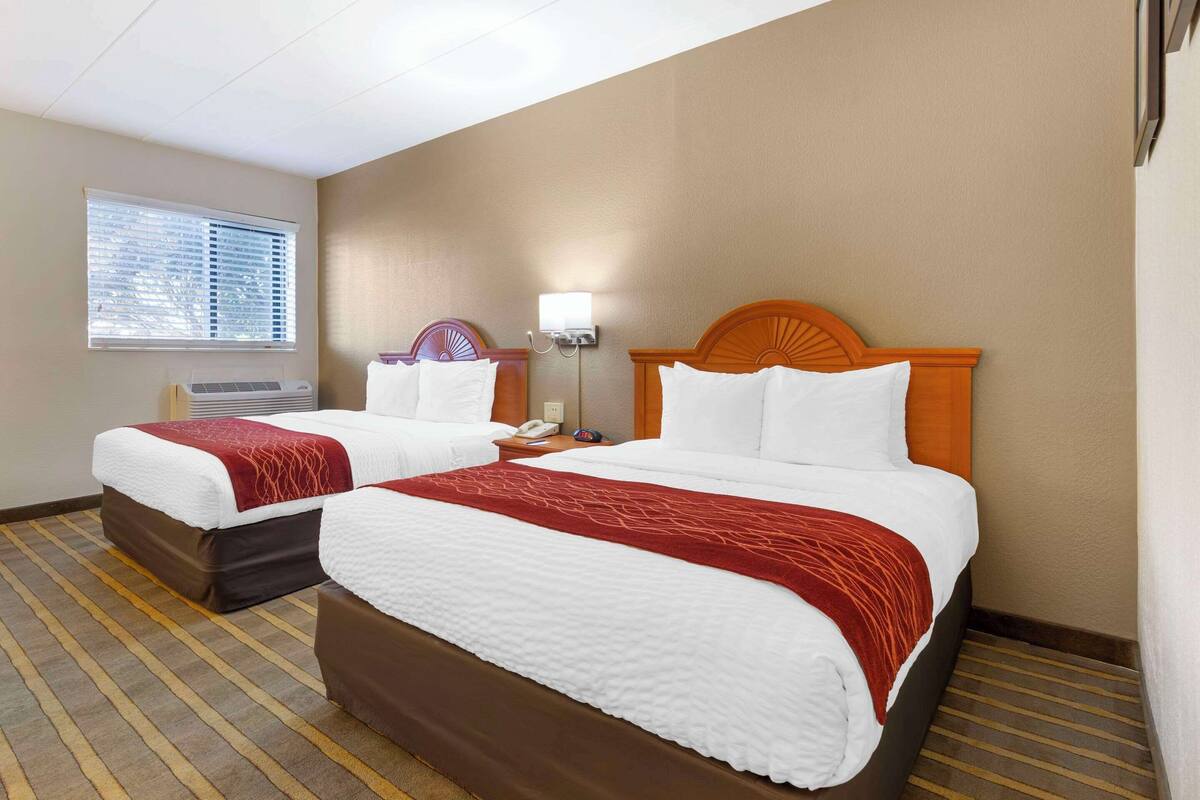 A hotel room with two beds, a window with blinds, an air conditioning unit, a nightstand with a phone, and a wall-mounted lamp.