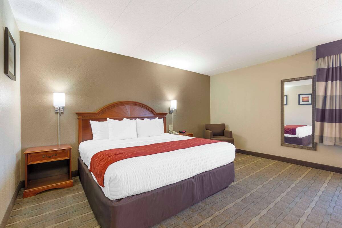 A hotel room with a large bed, a nightstand, a chair, and a mirror on the wall.
