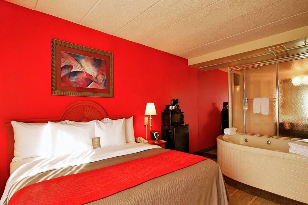 A hotel room with a bed, a red wall, a painting, a desk with a lamp, a coffee machine, and a bathtub.