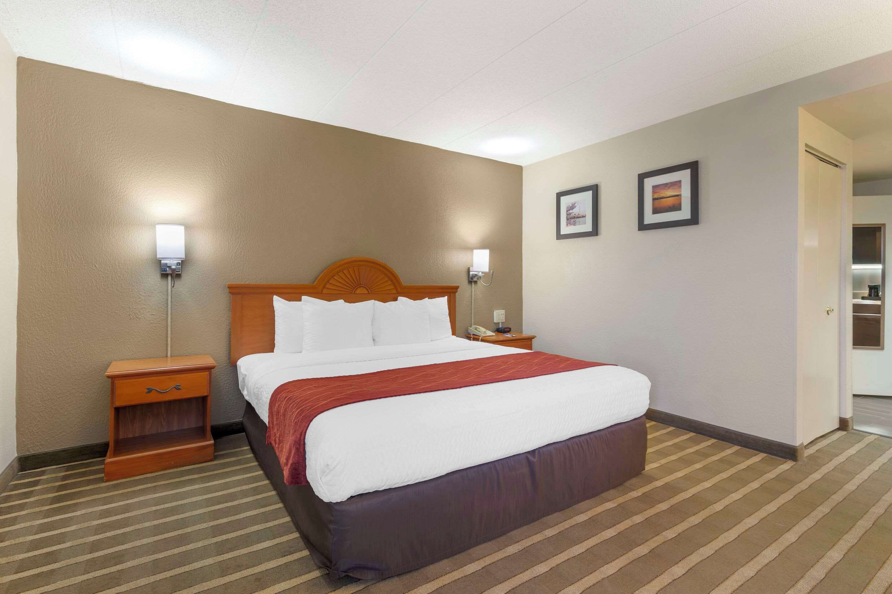Una habitación de hotel con una cama grande, cabecera de madera, mesita de noche, lámpara y dos cuadros en la pared.
