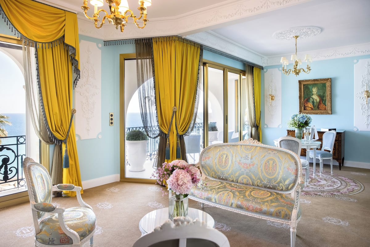 A room with a patterned sofa, a chair, a table with a vase of flowers, and a view of the sea through large windows with yellow curtains.