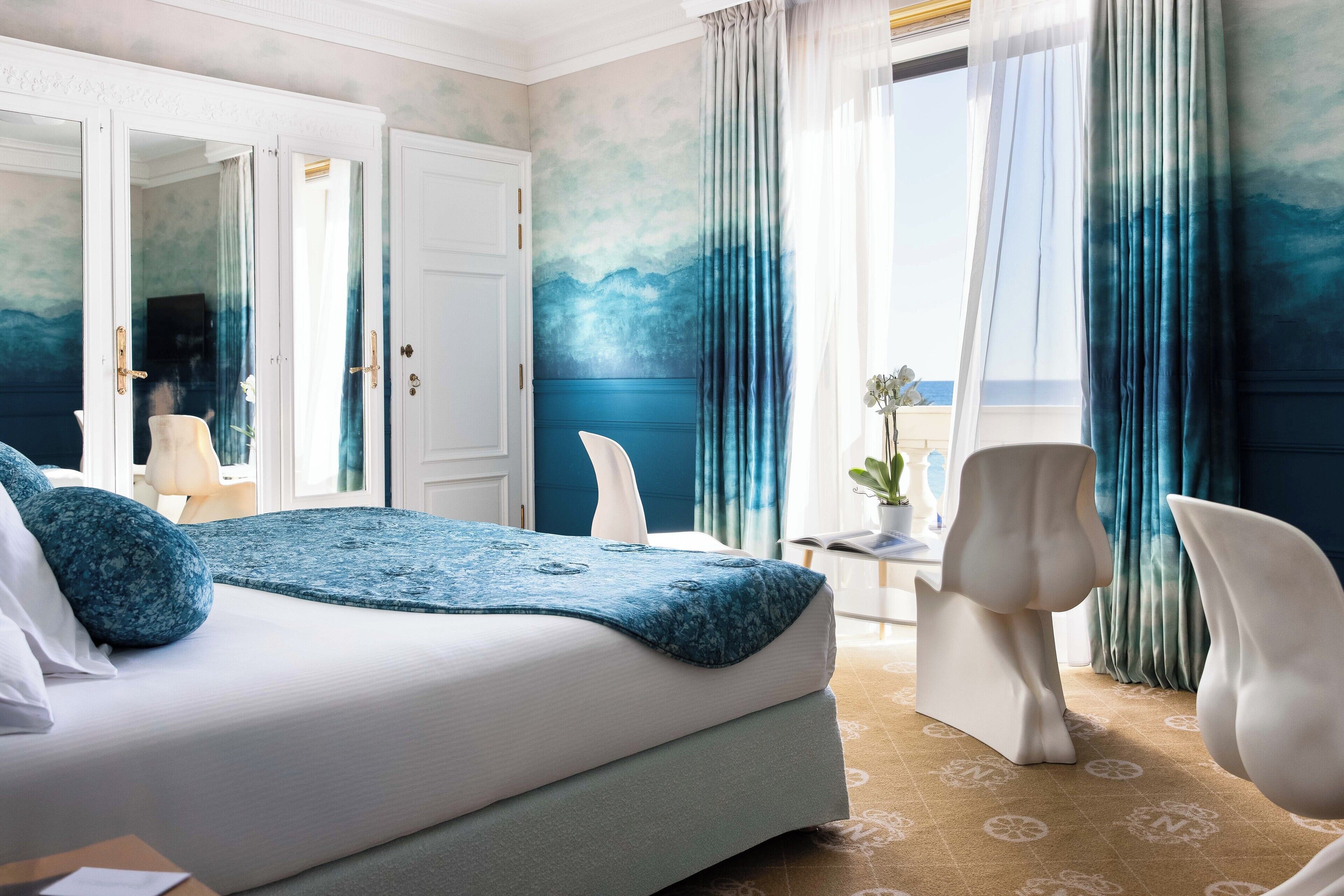 A hotel room with a bed, two chairs, a small table, and a view of the sea through the window.