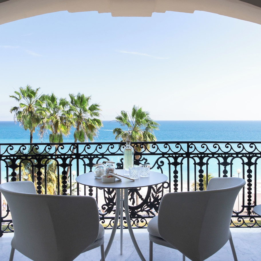 A balcony with a view of the sea, two white chairs, a small table with a vase and a bowl, and a potted plant.