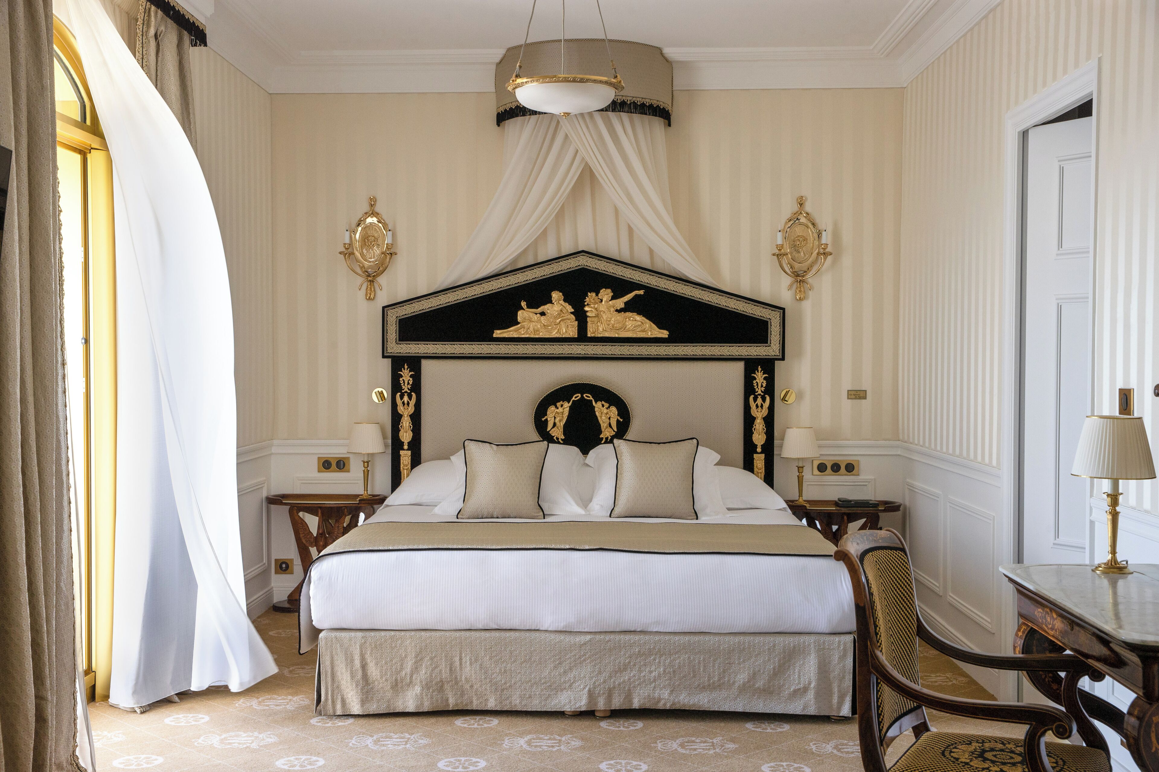 A large bed with a canopy, two bedside tables, a chair, and a desk with a lamp.