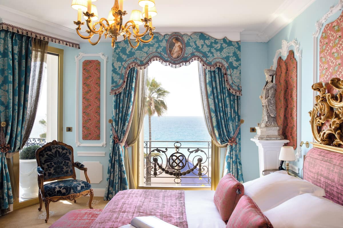 A bedroom with a large bed, a chair, a balcony with a view of the sea, and ornate decor.