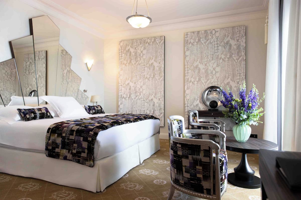 A hotel room with a large bed, a desk with a chair, a mirror, and a vase with flowers.