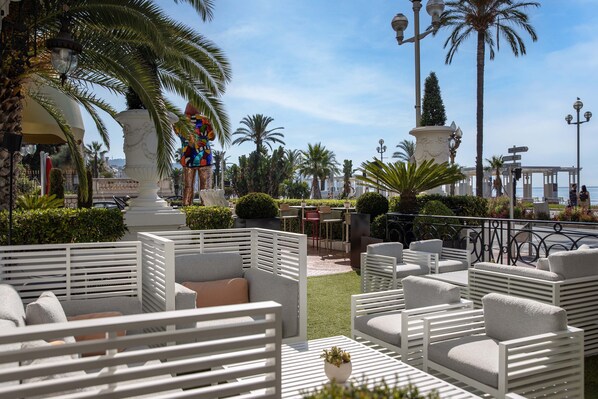 Un espacio al aire libre con mobiliario blanco, palmeras y una fuente decorativa.