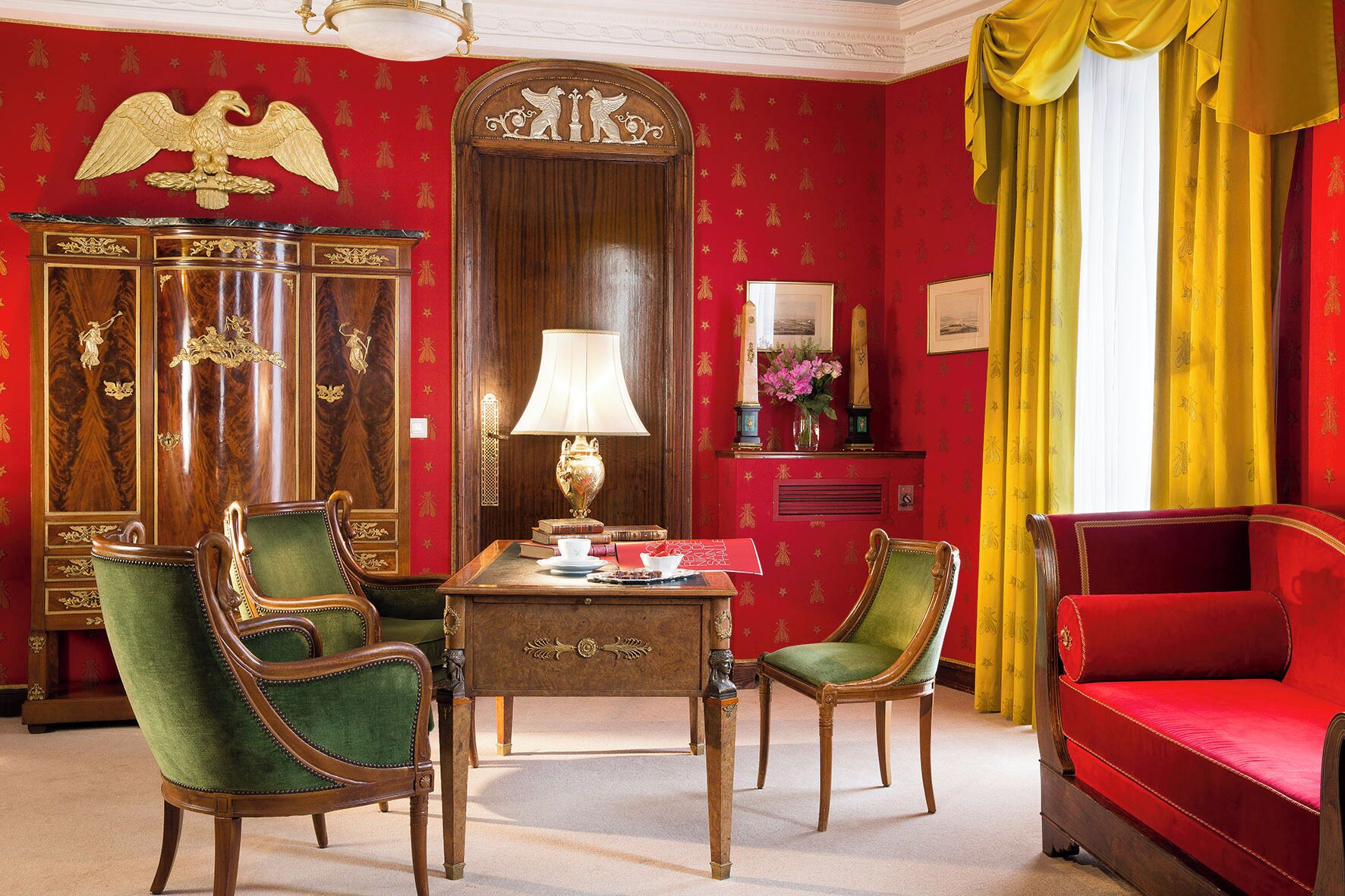 A room with a red wall, a wooden cabinet with gold detailing, a green upholstered chair, a red sofa, and a table with a lamp.