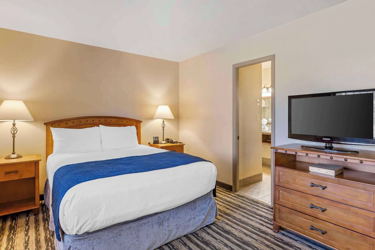 Habitación de hotel con cama, dos lámparas de noche, un televisor sobre un aparador de madera y una puerta que da a un baño.
