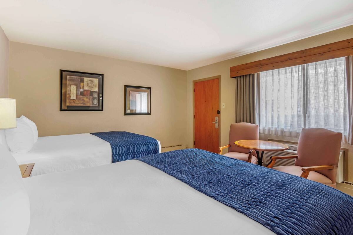 Una habitación de hotel con dos camas, una mesita, una silla y un ventanal con cortinas.