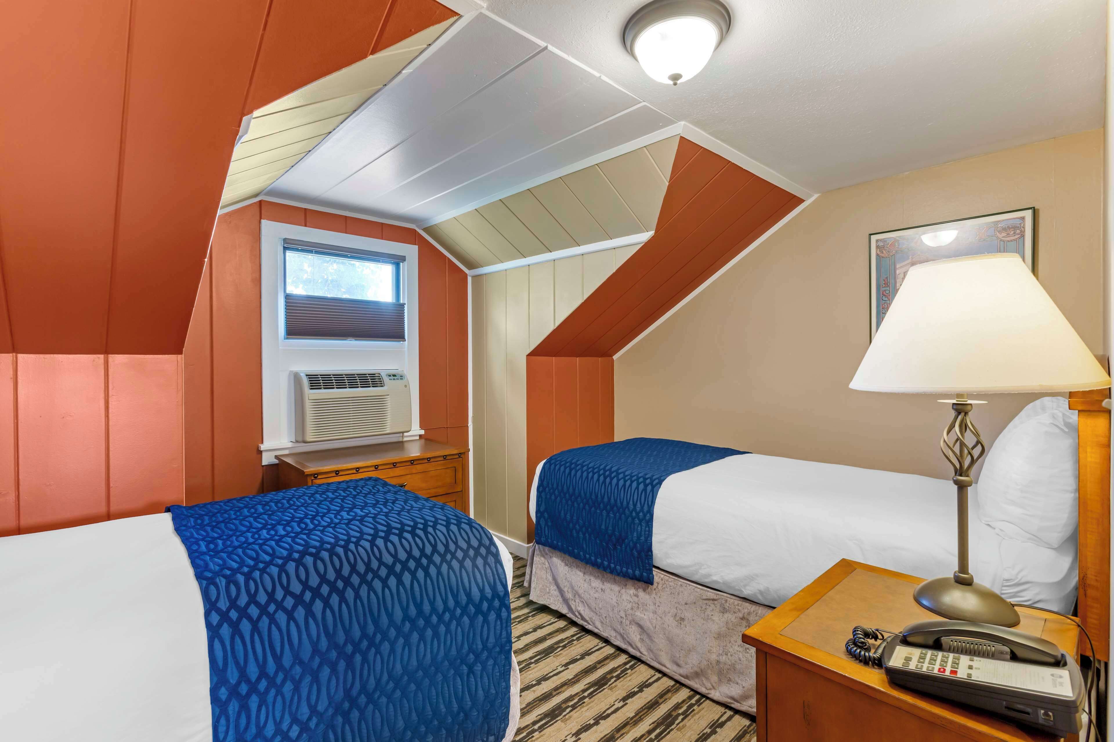 A hotel room with two beds, a desk with a lamp, a phone, and a window with blinds.