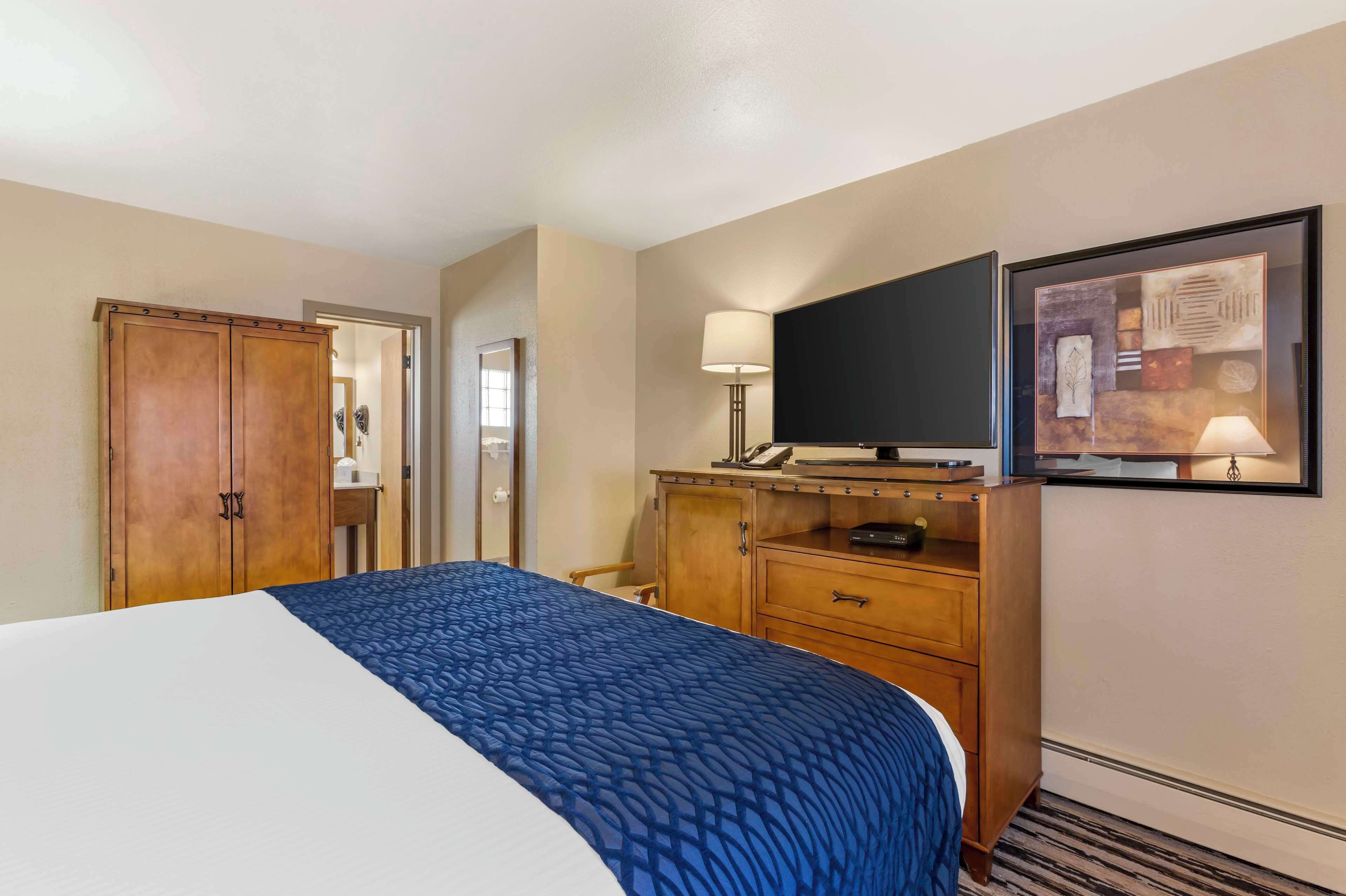 A hotel room with a large bed, wooden wardrobe, desk with a lamp, and a framed picture on the wall.