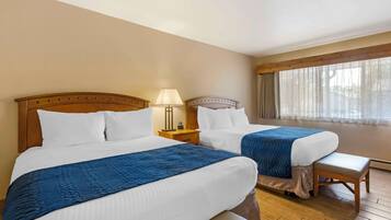 A hotel room with two beds, wooden headboards, a bench, a nightstand with a lamp, and a large window with sheer curtains.