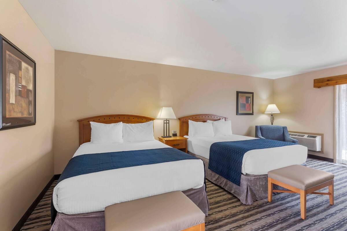 Una habitación de hotel con dos camas, un banco, una mesita de noche y un ventanal con cortinas.