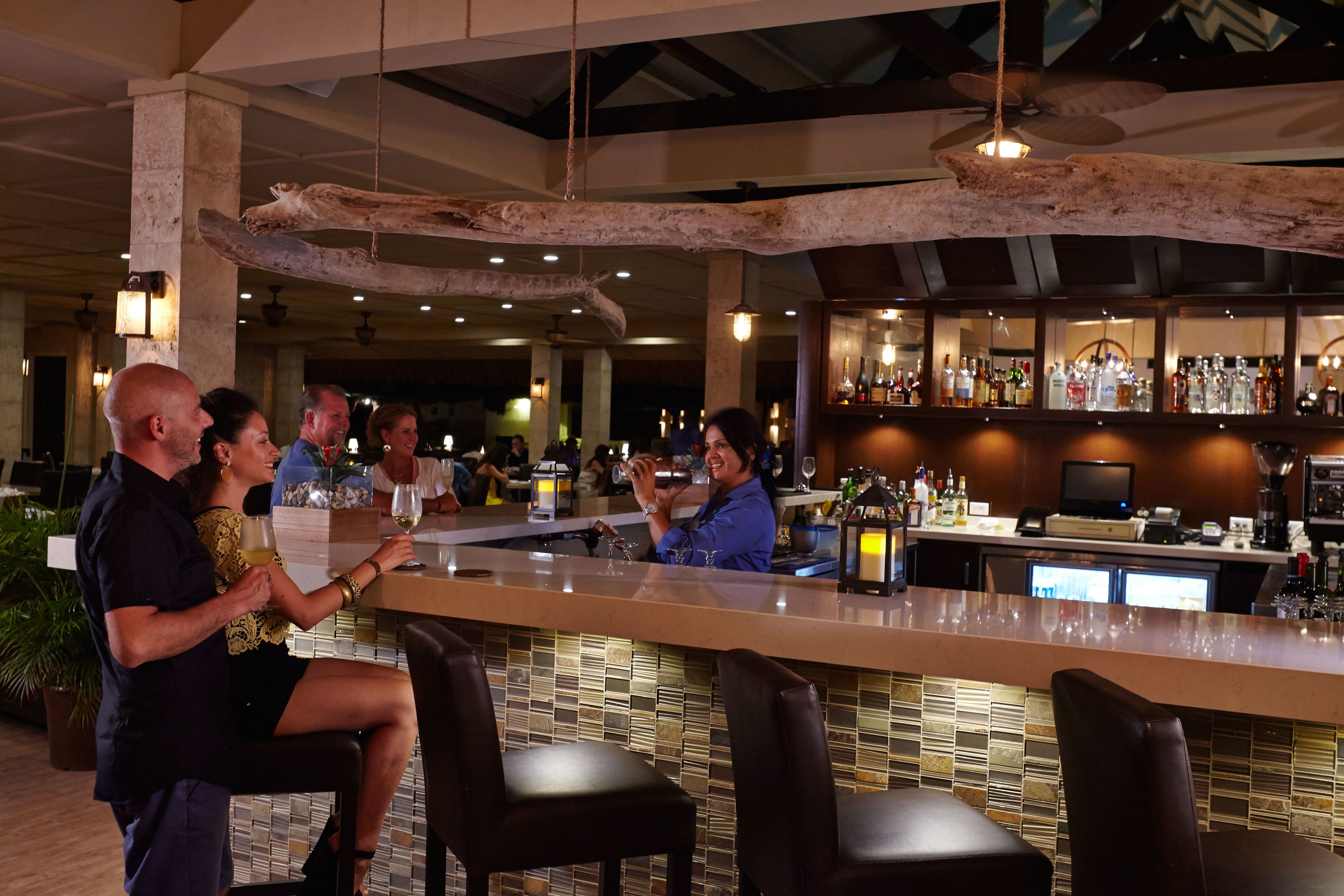 Una zona de bar con una barra alargada, diversas opciones de asientos y una amplia variedad de botellas y copas en estanterías.