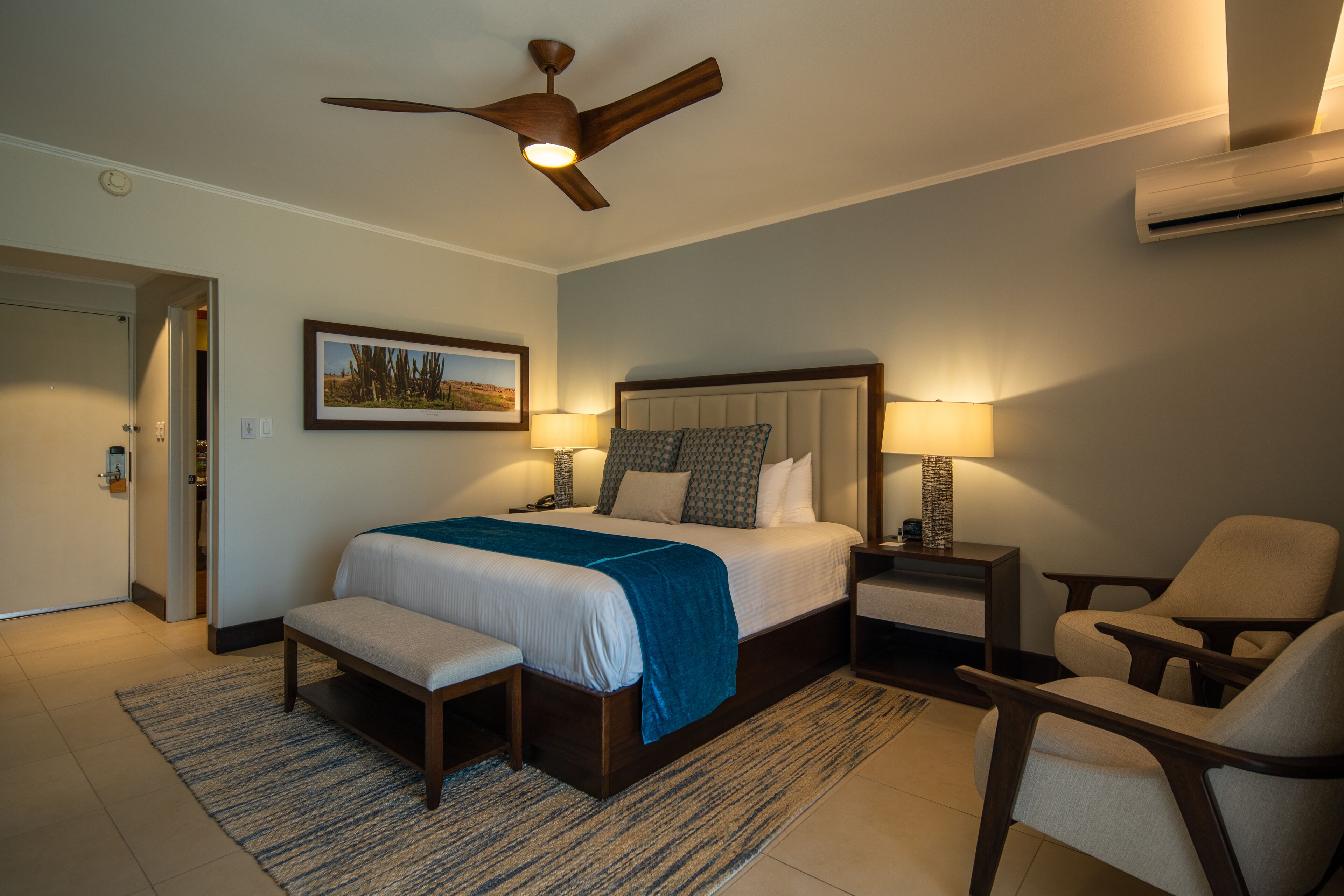 A hotel room with a bed, bedside tables, a bench, a chair, a ceiling fan, and an air conditioning unit.