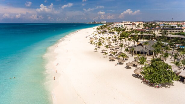 Una playa con aguas turquesas cristalinas, arena blanca y palmeras. Se ven edificios de varios pisos y algunas personas en la playa.