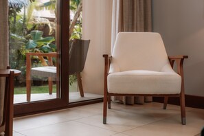 A room with a large sliding glass door leading to a garden, a white cushioned chair, and a wooden table.