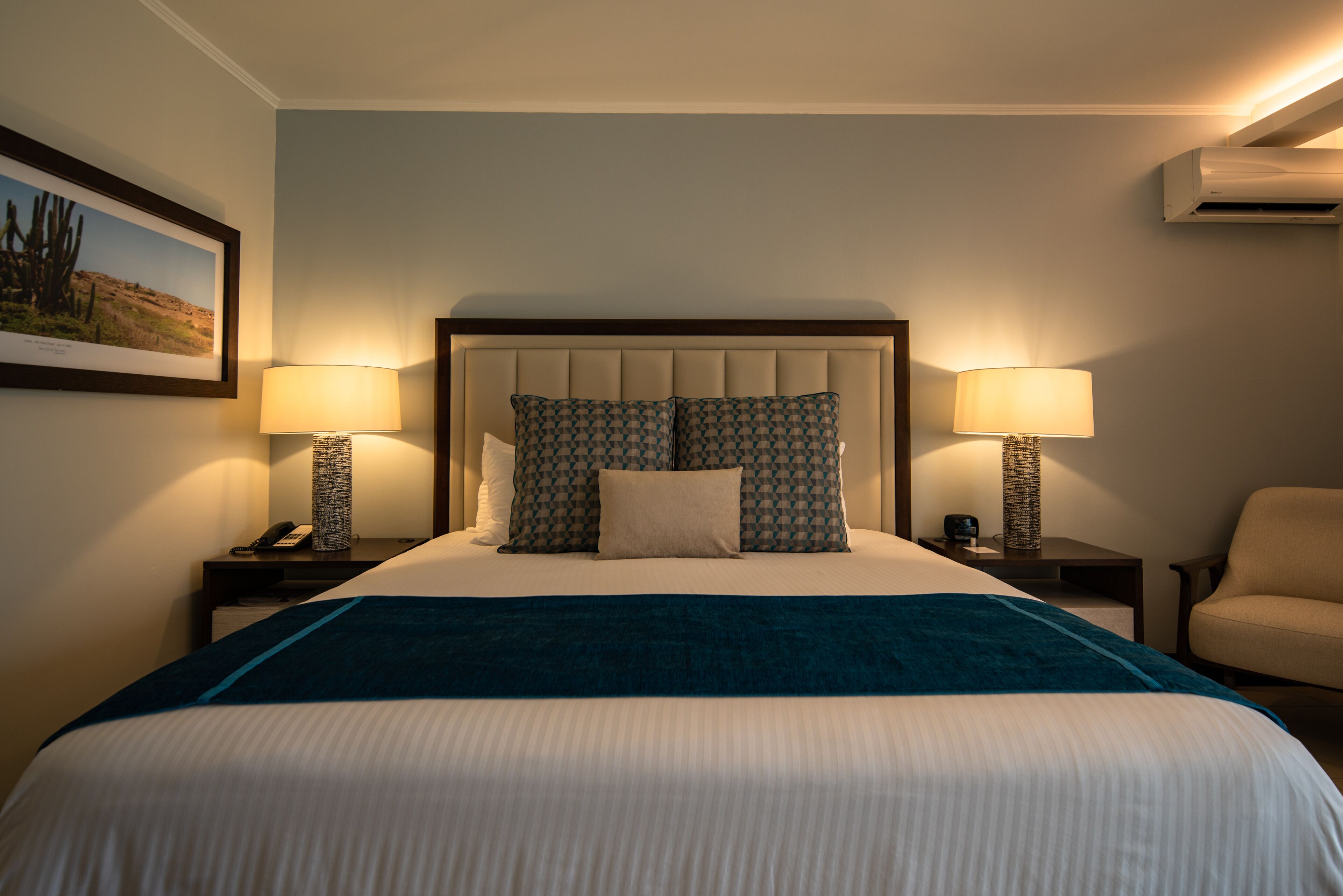 A neatly made bed with a blue blanket, two bedside lamps, a patterned pillow, and a framed picture on the wall.