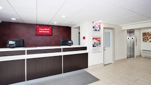Uma área de recepção com tela digital exibindo 'Wi-Fi Gratuito em Todos os Quartos', balcão de atendimento, interfone de segurança e elevador.