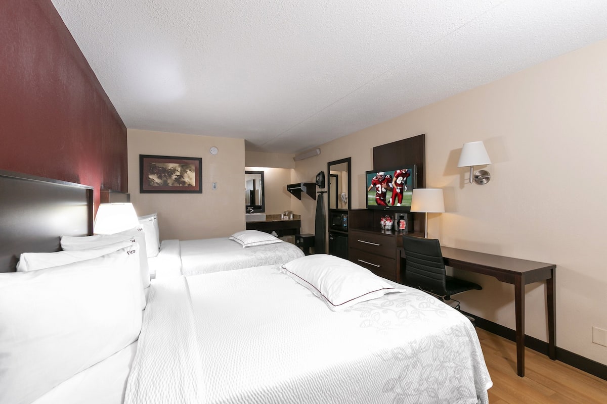 A hotel room with two beds, a desk, a chair, a TV, and a lamp.