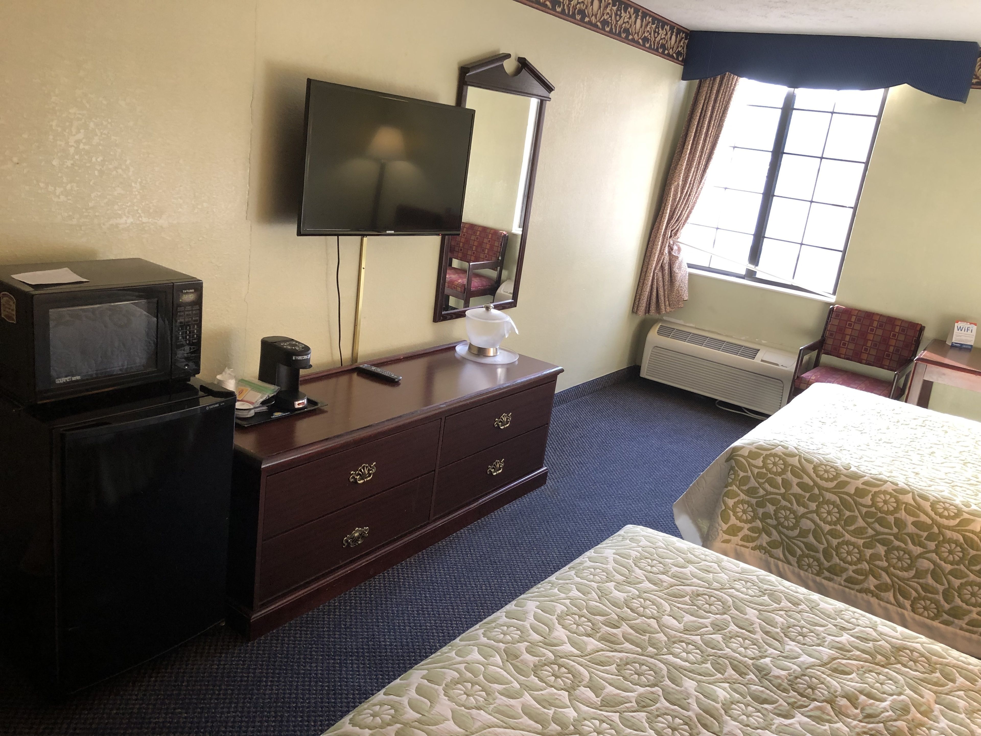 A hotel room with a TV, a desk with a lamp, a mirror, a chair, and two beds.