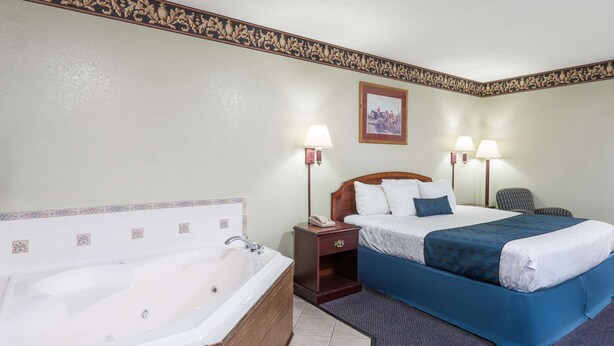 Habitación de hotel con una bañera grande, una cama con ropa de cama azul, dos lámparas de noche, una mesita de noche y un cuadro en la pared.