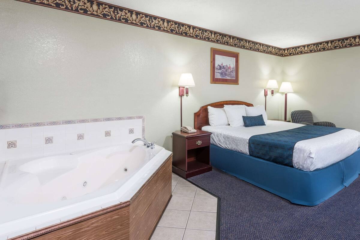 Une chambre d’hôtel avec une grande baignoire, un lit avec une literie bleue, deux lampes de chevet, une table de chevet et un tableau encadré au mur.