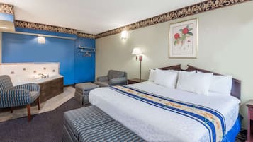 A hotel room with a large bed, two armchairs, a small table, a bathtub, and a wall-mounted TV.