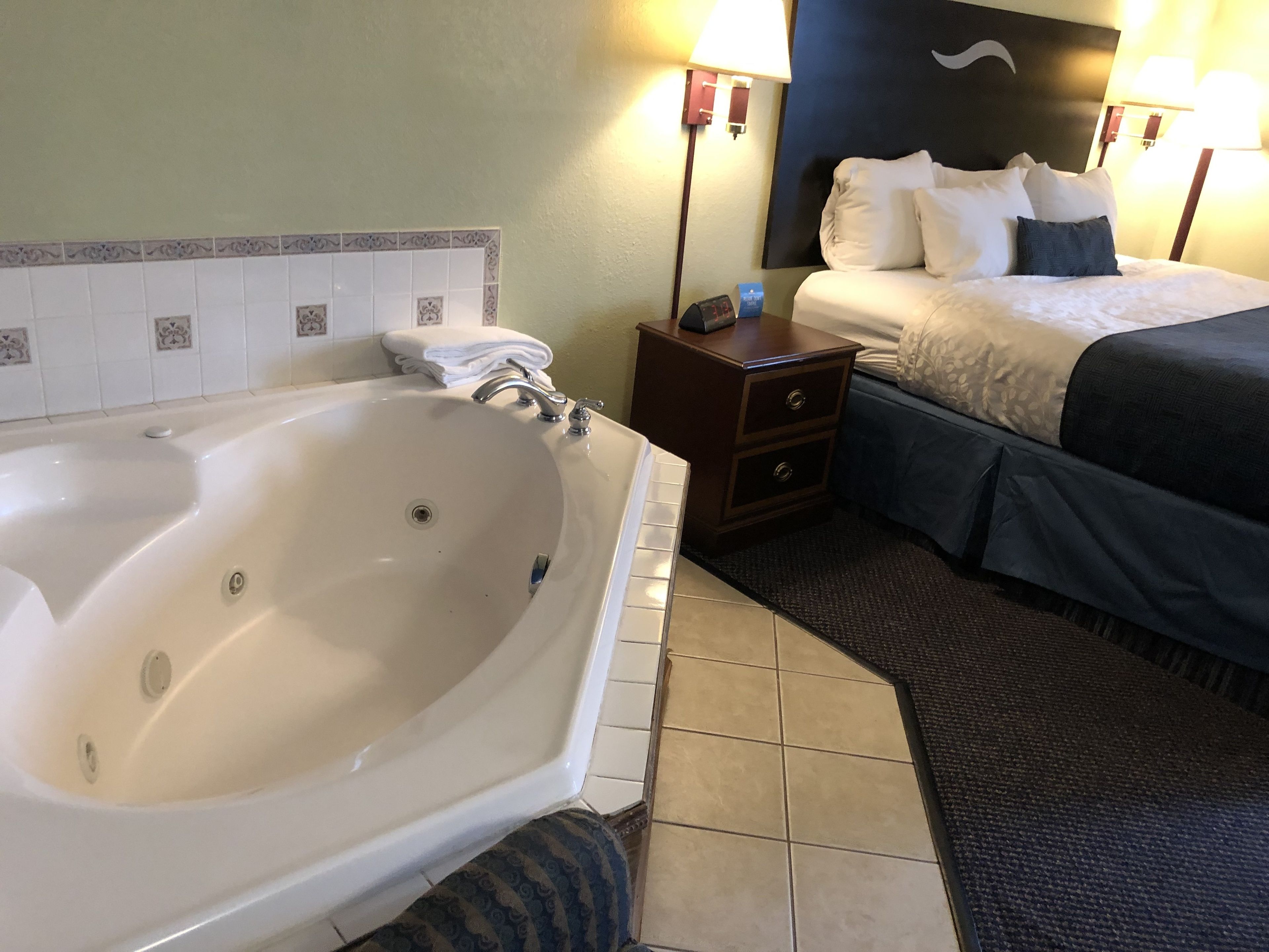 A hotel room with a large bathtub, a bed with a dark blue comforter, and a small wooden nightstand with a lamp.