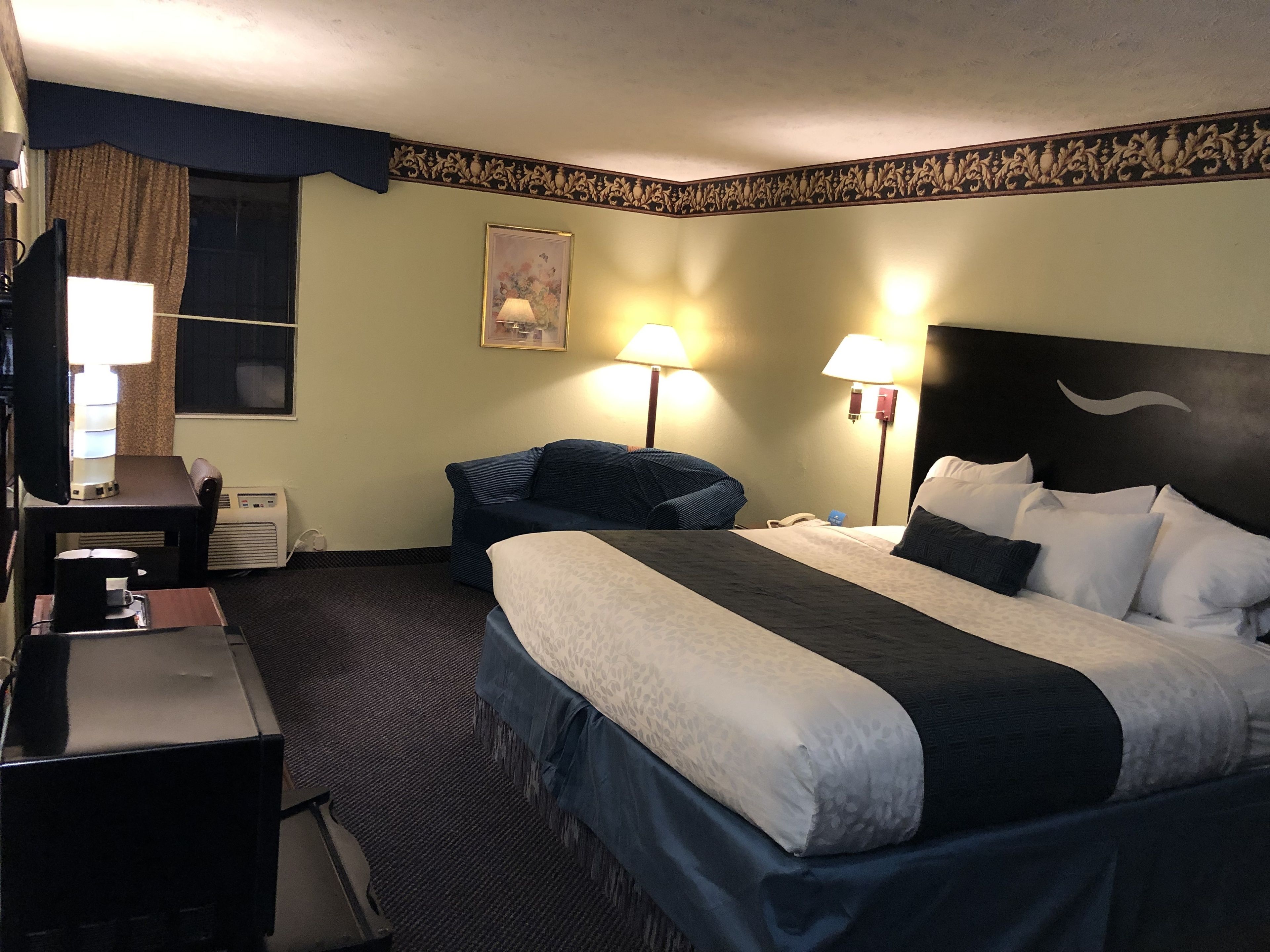A hotel room with a bed, bedside tables, a desk with a computer, a chair, a lamp, and a painting on the wall.