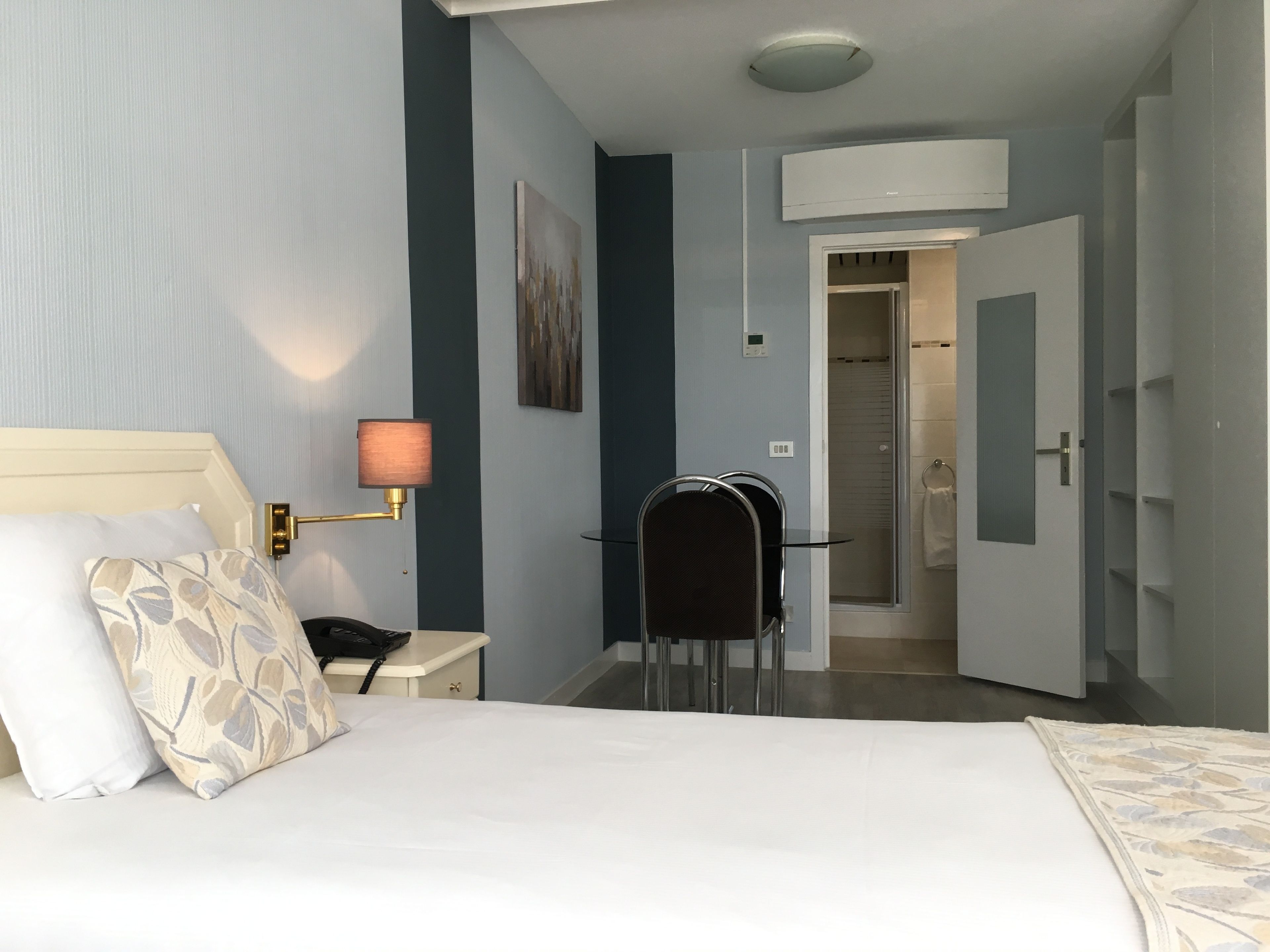 A hotel room with a bed, a desk, a chair, a lamp, a shower area, and a wardrobe.