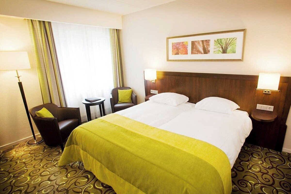 Una habitación de hotel con una cama grande, dos mesitas de noche, una silla, un escritorio y una ventana con cortinas.