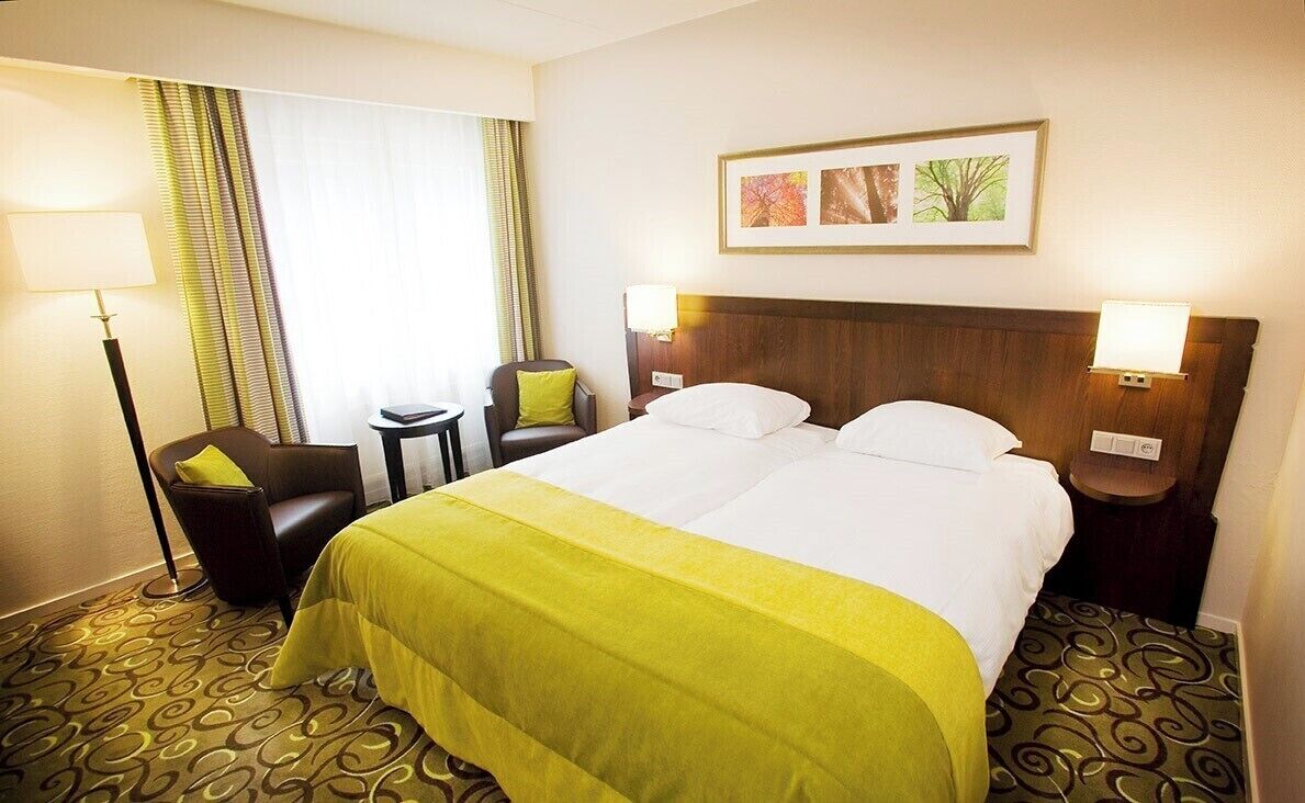 Habitación de hotel con cama doble, dos mesitas de noche, una silla, un escritorio, una lámpara, una ventana con cortinas y un cuadro en la pared.